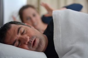 a middle-aged man asleep and snoring while his wife acts frustrated in the background